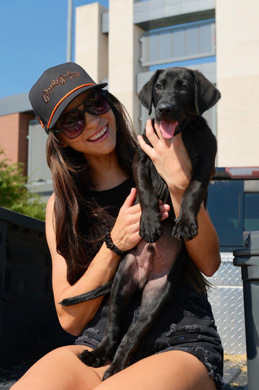 Woman wearing the Classic Cowboy Boone black and orange snapback while holding a black lab puppy.